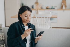 Smiling female holding bank credit card, smartphone, using online banking service at home. E-banking